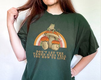 May include: Dark green t-shirt featuring a whimsical design. The graphic includes mushrooms, a frog, and a rainbow. The text on the shirt reads "DON'T LET THE TAMED ONES TELL YOU HOW TO LIVE".