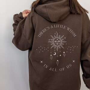 Puede incluir: Una sudadera con capucha marr&oacute;n con el texto "THERE'S A LITTLE WITCH IN ALL OF US" en blanco. El dise&ntilde;o incluye un sol, estrellas, lunas y cristales. La sudadera es de estilo jersey con capucha.