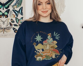 May include: Navy blue sweatshirt with a whimsical illustration of mushrooms, a fairy, and the text "believe in magic." The design features a star and other botanical elements. The sweatshirt is worn by a person.