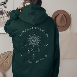 Puede incluir: Sudadera con capucha verde oscuro con el texto blanco "THERE'S A LITTLE WITCH IN ALL OF US". El dise&ntilde;o presenta un sol con una cara, estrellas y cristales colgantes. La sudadera tiene capucha y mangas largas.