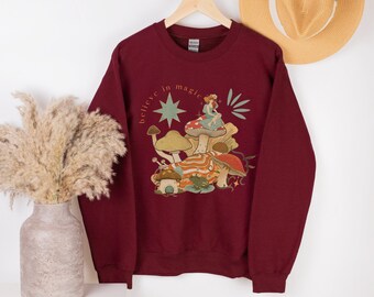 Puede incluir: Una sudadera granate con una ilustración caprichosa que presenta setas, un hada y el texto "believe in magic". El diseño incluye una estrella y otros elementos botánicos. La sudadera está colgada en una percha de madera.