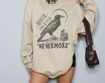 Könnte beinhalten: Beiges Sweatshirt mit schwarzer Grafik eines Raben, Mond und dem Text "QUOTH THE RAVEN NEVERMORE". Das langärmlige Oberteil ist im lässigen Stil gehalten.