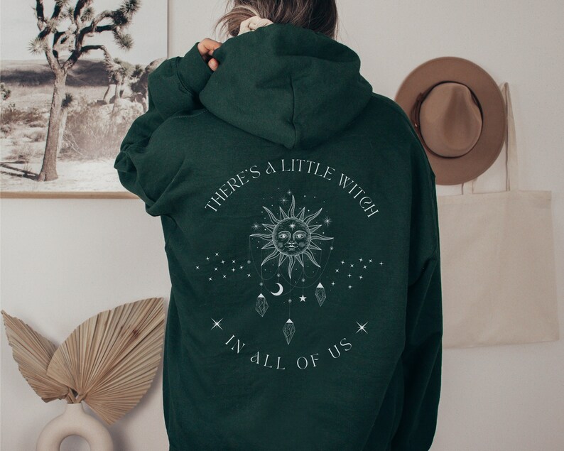 Puede incluir: Sudadera con capucha verde oscuro con el texto blanco "THERE'S A LITTLE WITCH IN ALL OF US". El dise&ntilde;o presenta un sol con una cara, estrellas y cristales colgantes. La sudadera tiene capucha y mangas largas.
