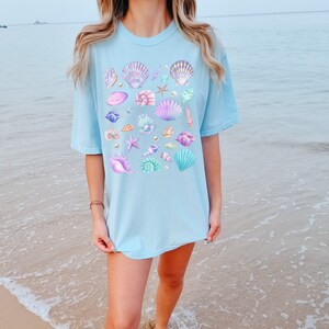 Op de afbeelding: Lichtblauw T-shirt met een aquarelontwerp van schelpen in verschillende kleuren, waaronder paars, roze en teal. Het ontwerp bevat ook zeesterren en parels. Het shirt heeft een relaxte, oversized pasvorm.