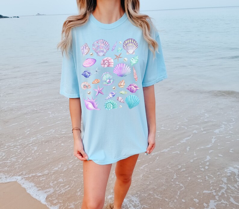 Op de afbeelding: Lichtblauw T-shirt met een aquarelontwerp van schelpen in verschillende kleuren, waaronder paars, roze en teal. Het ontwerp bevat ook zeesterren en parels. Het shirt heeft een relaxte, oversized pasvorm.