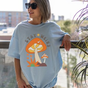 May include: Light blue t-shirt with a retro mushroom graphic and the text "STAY WEIRD". The design features an orange mushroom with a white stem, a smaller mushroom, and orange flowers. The shirt is a comfortable, casual style.