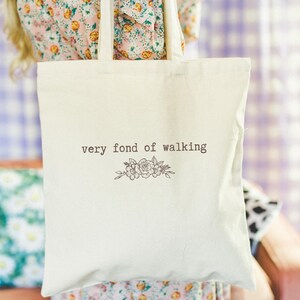May include: Off-white tote bag featuring the phrase "very fond of walking" and a floral illustration. The bag is held up by a person. The background is a blurred mix of colors.