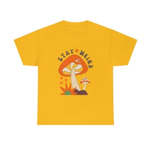May include: Mustard yellow t-shirt with a retro-style mushroom graphic. The design features a large orange mushroom with white spots, a smaller mushroom, and the text "STAY WEIRD" in a circular arrangement.