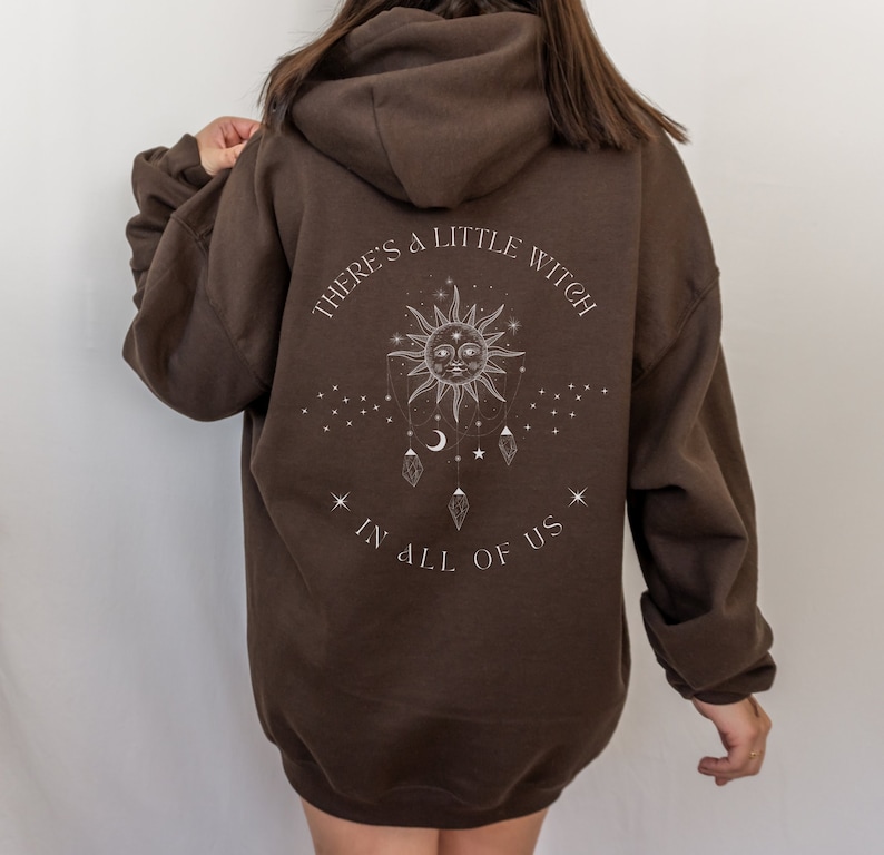 Puede incluir: Una sudadera con capucha marr&oacute;n con el texto "THERE'S A LITTLE WITCH IN ALL OF US" en blanco. El dise&ntilde;o incluye un sol, estrellas, lunas y cristales. La sudadera es de estilo jersey con capucha.