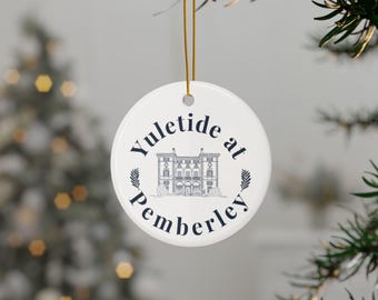 May include: A white, round Christmas ornament with the text "Yuletide at Pemberley" in navy blue script. A detailed illustration of a stately building is centered on the ornament, with sprigs of greenery on either side. The ornament hangs from a gold string.