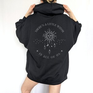 Puede incluir: Sudadera con capucha negra con mangas largas. La parte trasera presenta un gr&aacute;fico con un sol, una luna, estrellas y el texto "THERE'S A LITTLE WITCH IN ALL OF US."