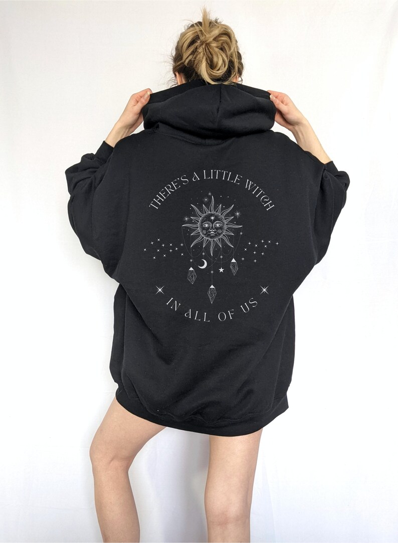 Puede incluir: Sudadera con capucha negra con mangas largas. La parte trasera presenta un gr&aacute;fico con un sol, una luna, estrellas y el texto "THERE'S A LITTLE WITCH IN ALL OF US."