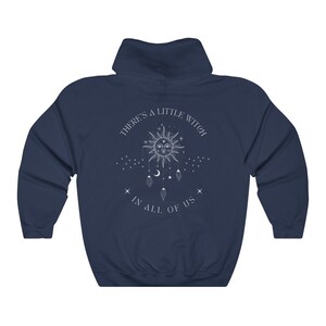 Puede incluir: Sudadera con capucha azul marino con un gr&aacute;fico blanco en la espalda. El gr&aacute;fico presenta un sol, estrellas y el texto "THERE'S A LITTLE WITCH IN ALL OF US."