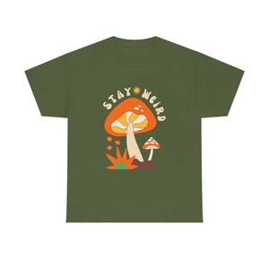 May include: Olive green t-shirt featuring a retro-style mushroom graphic with the text "Stay Weird." The design includes an orange mushroom with a yellow stem, small orange flowers, and a smaller mushroom. A fun and whimsical design.