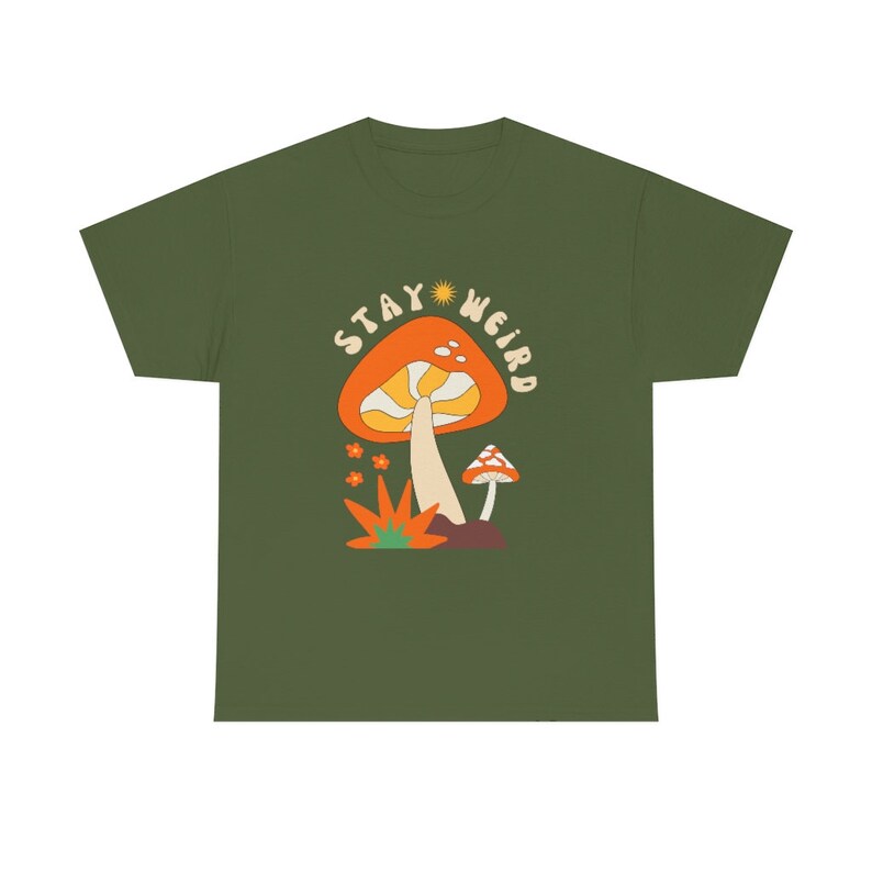 May include: Olive green t-shirt featuring a retro-style mushroom graphic with the text "Stay Weird." The design includes an orange mushroom with a yellow stem, small orange flowers, and a smaller mushroom. A fun and whimsical design.