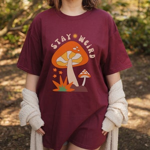 May include: Burgundy t-shirt with a mushroom graphic and the text "STAY WEIRD". The graphic features an orange mushroom with a cream-colored stem and a smaller mushroom. The shirt is a relaxed fit.
