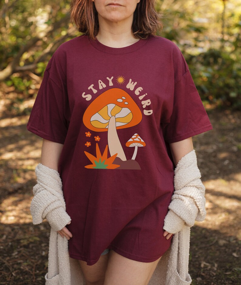 May include: Burgundy t-shirt with a mushroom graphic and the text "STAY WEIRD". The graphic features an orange mushroom with a cream-colored stem and a smaller mushroom. The shirt is a relaxed fit.