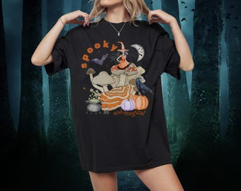 May include: Black t-shirt with a colorful graphic featuring a witch on a mushroom, a crescent moon, and the words "spooky and magical." The design includes pumpkins, a cauldron, and a raven, perfect for Halloween.