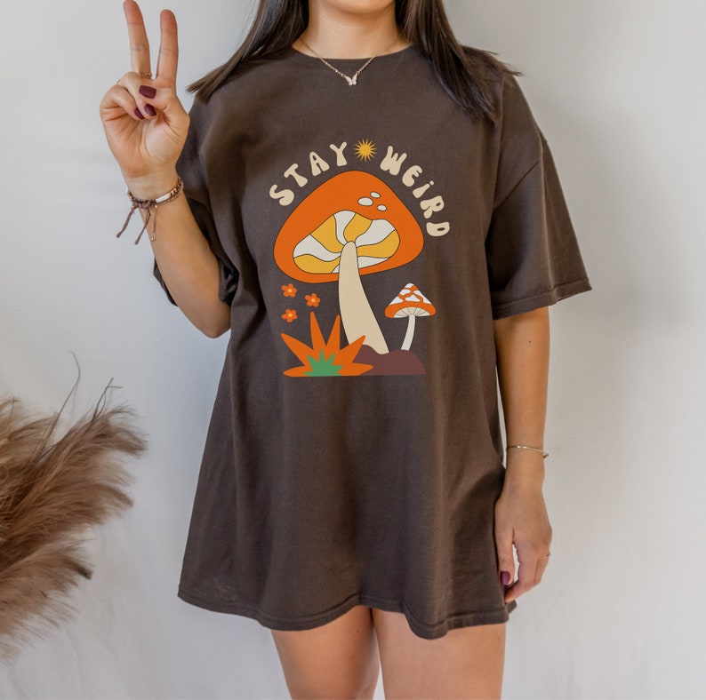May include: A dark brown t-shirt featuring a colorful mushroom graphic with the text "STAY WEIRD". The design includes an orange mushroom with yellow and white details, smaller mushrooms, and floral accents. The shirt is a relaxed fit.