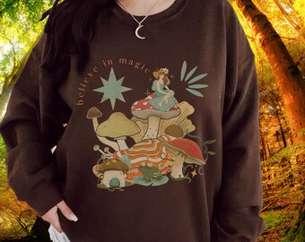 Puede incluir: Una sudadera marrón oscuro con un gráfico caprichoso que presenta setas, un hada y el texto "believe in magic" en un arco circular. El diseño incluye una estrella y otros elementos botánicos. La sudadera es de color liso.