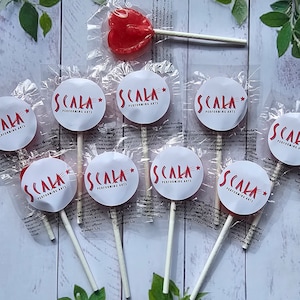 Può includere: Nove lecca-lecca rotondi bianchi con una stella rossa e il testo "SCALA PERFORMING ARTS" in rosso. I lecca-lecca sono avvolti in plastica trasparente e hanno bastoncini bianchi. Un lecca-lecca rosso a forma di cuore è sullo sfondo.