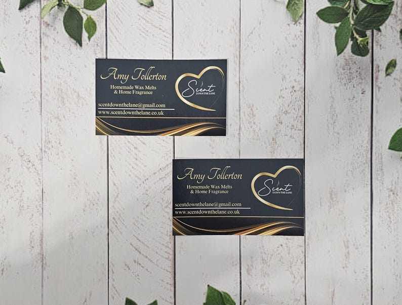 May include: Two black business cards with gold lettering and a gold heart logo. The text on the cards reads "Amy Tollerton", "Homemade Wax Melts & Home Fragrance", "scentdownthelane@gmail.com", and "www.scentdownthelane.co.uk". The logo is a gold heart with the word "Scent" inside.
