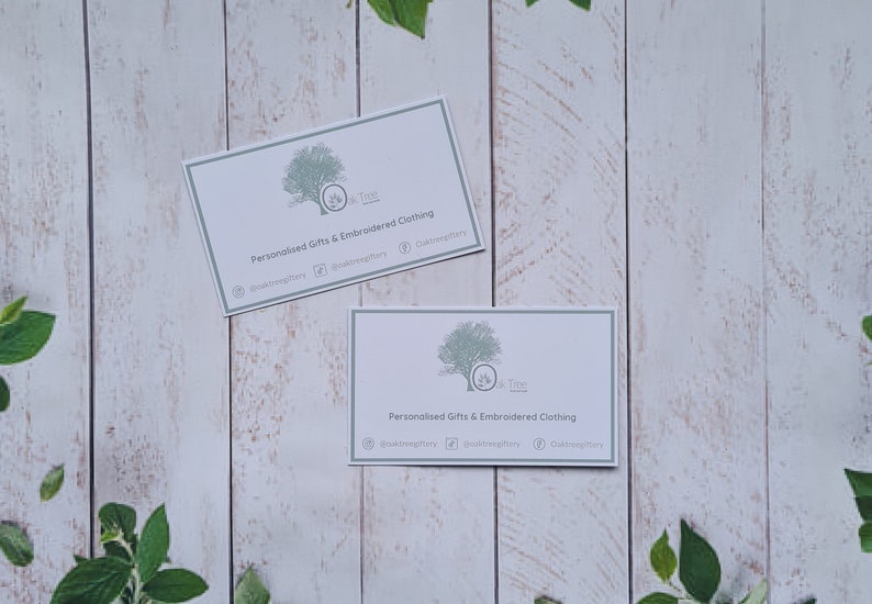 May include: Two white business cards with a green tree logo and the text "Oak Tree" and "Personalized Gifts & Embroidered Clothing" on a white wood background.
