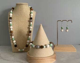 May include: A jewelry set consisting of a beaded necklace, a beaded bracelet, and a pair of drop earrings. The pieces feature a repeating pattern of white, light green, brown, and grey spherical beads separated by small silver-toned metal spacers.