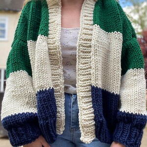 Puede incluir: Un cárdigan de punto grueso con un patrón de bloques de color verde, blanco y azul marino. El cárdigan tiene un corte holgado y mangas largas.