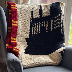 May include: A crocheted blanket featuring a black silhouette of Hogwarts castle on a beige background. The blanket has a red and gold border.