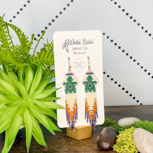 Puede incluir: Un par de pendientes de cuentas verdes, blancas, naranjas y azules con cierres de oro rellenos. Los pendientes se muestran en una tarjeta blanca con el texto "Wabi Sabi Bead Co Handmade 14k gold filled ear wires".