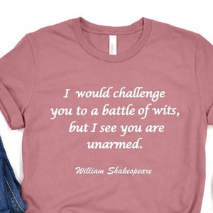 May include: Dusty rose t-shirt with the quote "I would challenge you to a battle of wits, but I see you are unarmed." by William Shakespeare. The shirt is paired with blue jeans and white slip-on shoes.