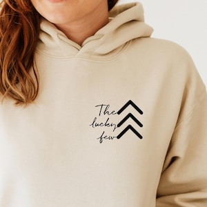 May include: A beige hoodie with a black graphic that says "The lucky few" with three black arrows pointing upwards.