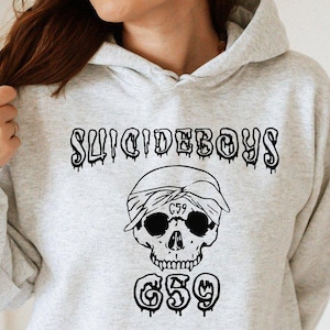 May include: Light gray hoodie with a black graphic featuring the text "SUICIDEBOYS" above a skull wearing a bandana and sunglasses, with "G59" below. The hoodie has a drawstring hood and a front pocket.
