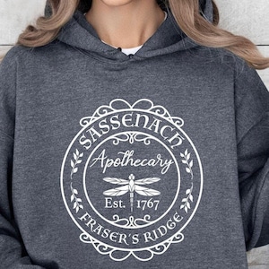 May include: Dark gray hoodie with a white circular design featuring the text "Sassenach Apothecary Fraser's Ridge Est. 1767" and a dragonfly illustration. The hoodie has a drawstring hood and a front pocket.