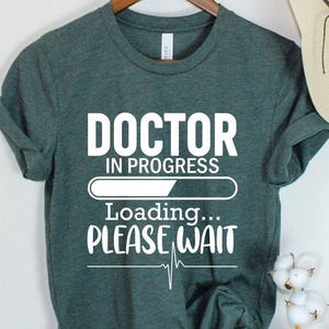 May include: A dark green t-shirt with the text "DOCTOR IN PROGRESS Loading... PLEASE WAIT" in white. A beige sun hat with a black bow hangs nearby. The shirt is displayed on a wooden hanger.