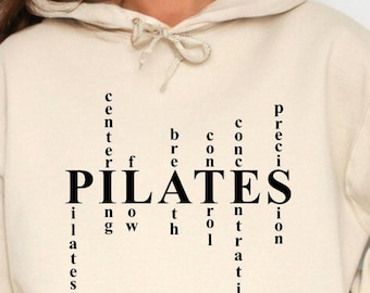 May include: A beige Pilates hoodie with a front pocket and drawstring hood. The word "PILATES" is printed in bold black letters, with related words like "center," "breath," and "precision" arranged vertically around it. A comfortable, casual style.
