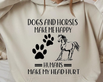 May include: Beige hoodie featuring the text "DOGS AND HORSES MAKE ME HAPPY" and "HUMANS MAKE MY HEAD HURT". The design includes black paw prints and a horse illustration. The hoodie has a drawstring hood and a front pocket.