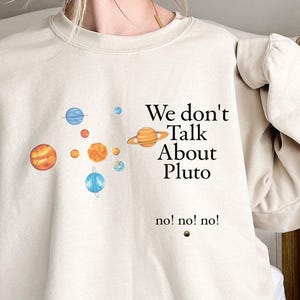 May include: Beige sweatshirt featuring a colorful planet graphic and the text "We don't Talk About Pluto" with "no! no! no!" below. The design includes illustrations of planets in shades of orange, blue, and yellow.