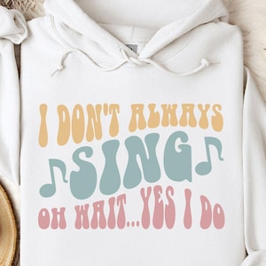 May include: White hoodie with the text "I DON'T ALWAYS SING OH WAIT...YES I DO" in retro-style lettering. The text is in shades of yellow, blue, and pink. The hoodie has a drawstring and a front pocket.