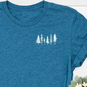 May include: Teal blue t-shirt with a small white tree graphic on the chest. The shirt has short sleeves and a crew neck. The shirt is paired with blue jeans and a small floral arrangement.