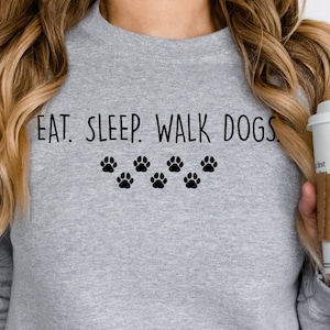 May include: A light gray sweatshirt with the text "EAT. SLEEP. WALK DOGS." in black, along with six black paw prints. The person is holding a white coffee cup with a brown sleeve.