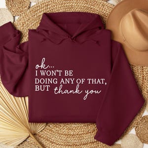 May include: A maroon hoodie with white text that reads "ok... I won't be doing any of that, but thank you." The hoodie is displayed on a woven surface with a brown hat in the background. The hoodie has a hood and long sleeves.