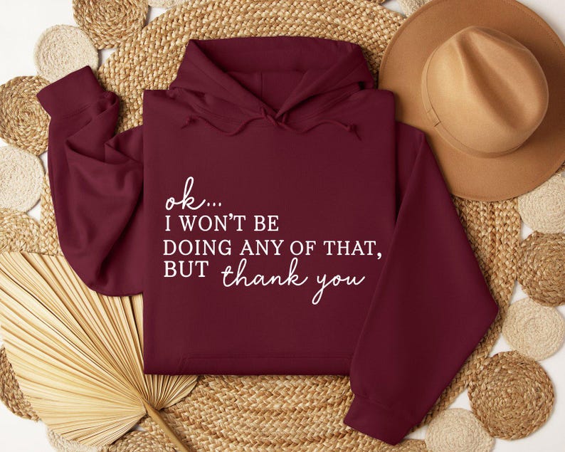 May include: A maroon hoodie with white text that reads "ok... I won't be doing any of that, but thank you." The hoodie is displayed on a woven surface with a brown hat in the background. The hoodie has a hood and long sleeves.
