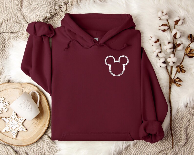 以下が含まれることがあります： 前面にミッキーマウスの頭の白いアウトラインが付いた、マルーン色のパーカー。 パーカーには、ドローストリングフードとフロントポケットが付いています。 画像は、木製のトレイ、マグカップ、雪の結晶の装飾が施された白い毛皮のような表面にあります。