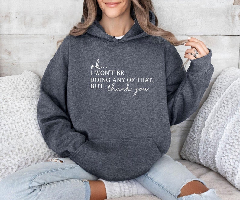 May include: A dark gray hoodie with a front pocket and a drawstring hood. The hoodie features the text "ok... I won't be doing any of that, but thank you" in white script. The sweatshirt is made of a soft material.