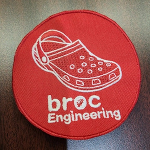 Könnte beinhalten: Roter gestickter Aufnäher mit einer weißen Umrandung eines Clogs und dem Text "broc Engineering" in weiß.