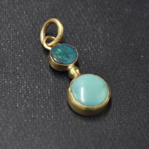 May include: A gold pendant featuring two gemstones: a round, light blue turquoise stone and an oval, dark teal opal. The pendant has a small loop at the top for a chain.