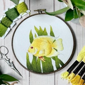 May include: An embroidered yellow fish with green leaves on a white background in a wooden embroidery hoop.