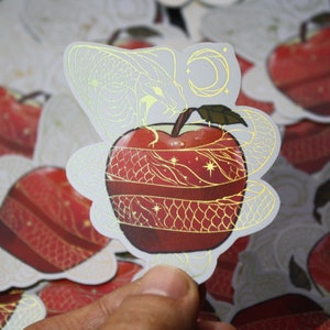Könnte beinhalten: Ein roter Apfel mit goldenen Folienverzierungen und einem Schlangenmuster. Der Apfel ist von einem weißen Hintergrund mit goldenen Sternen und einem Halbmond umgeben.